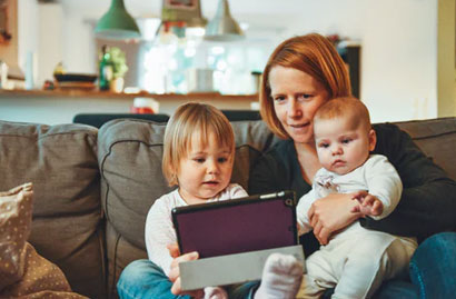 Two Blonde-Haired Babies Sitting in Their Mother's Lap Watching Something on a Table